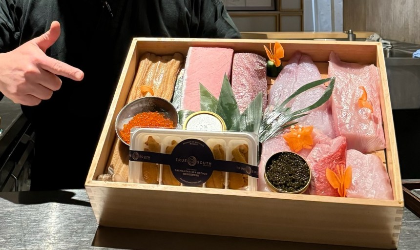 Hand points at wodden box filled with raw fish (nigiri)