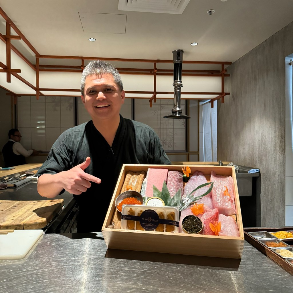 Chef points at sashimi nigiri in a wooden crate.